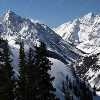 Snow-covered mountain peaks rise under a clear blue sky, with dark evergreen trees in the foreground. The landscape features deep valleys and dramatic slopes, creating a striking winter scene in a rugged alpine environment.