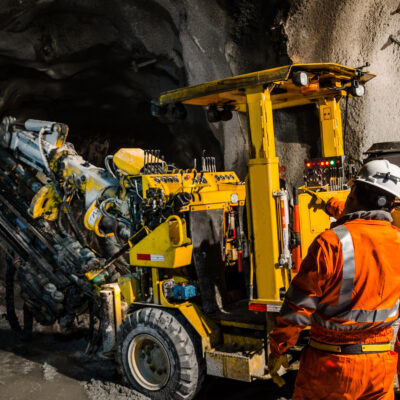 Two workers in orange safety suits and helmets operate a large yellow drilling machine inside an underground tunnel with rough, rocky walls.