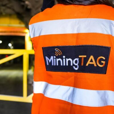 A person wearing an orange safety vest with "MiningTAG" on the back stands in front of a gated entrance to a mine tunnel, illuminated by yellow lights inside.