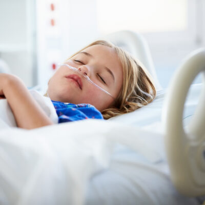 A young child with a nasal oxygen tube sleeps in a hospital bed, wearing a hospital gown. The child’s hands are folded on their chest and medical equipment is visible around the bed.