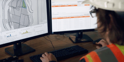 A person wearing a safety vest and hard hat is working at a desk with two computer monitors displaying maps, charts, and data tables.