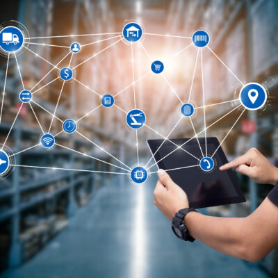 Person using a tablet in a warehouse with digital icons representing logistics and supply chain connections overlayed, symbolizing modern technology and inventory management.