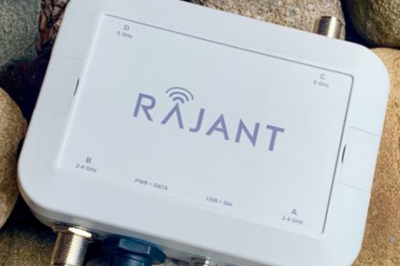 A white Rajant wireless device with four antenna ports sits on a bed of large rocks. The device has labeled ports and LED indicators on its front panel.