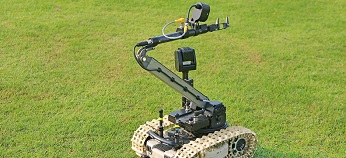 A tracked robot with a mechanical arm and camera is positioned on green grass. The arm is extended, and the robot appears to be designed for remote handling or inspection tasks.
