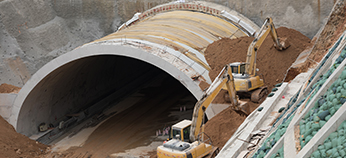 Two excavators are working near the entrance of a large tunnel under construction, moving dirt and shaping the landscape around the tunnel’s concrete structure.