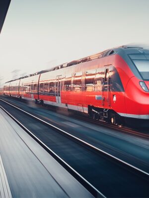 A bright red modern passenger train speeds through a blurred urban train station, creating a sense of motion and urgency as it travels along the tracks.