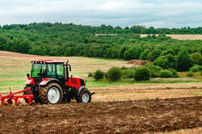 A red tractor plowing a brown field with green trees and hills in the background under a cloudy sky.