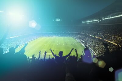 A large, brightly lit soccer stadium filled with cheering fans. People in the stands have their arms raised, celebrating as a match takes place on the green field under the lights.