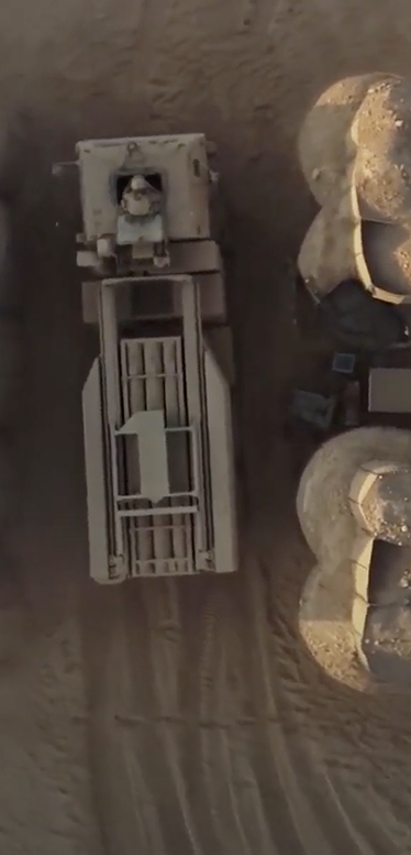 Aerial view of a military vehicle driving on a dirt road between large sand-colored barriers or structures in a desert environment.