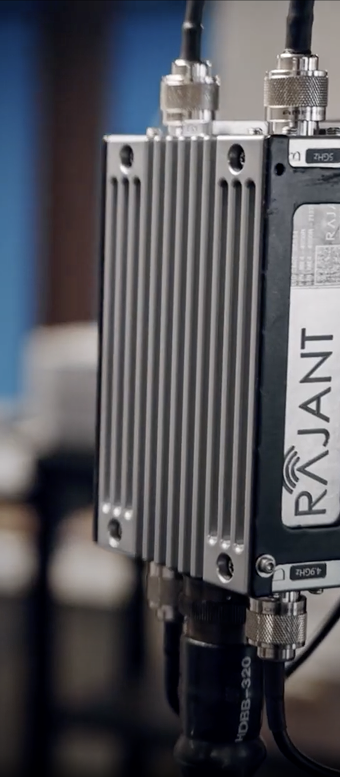 A close-up of a silver and black electronic device with large cooling fins, labeled "RAJANT" on the side, connected to several cables.