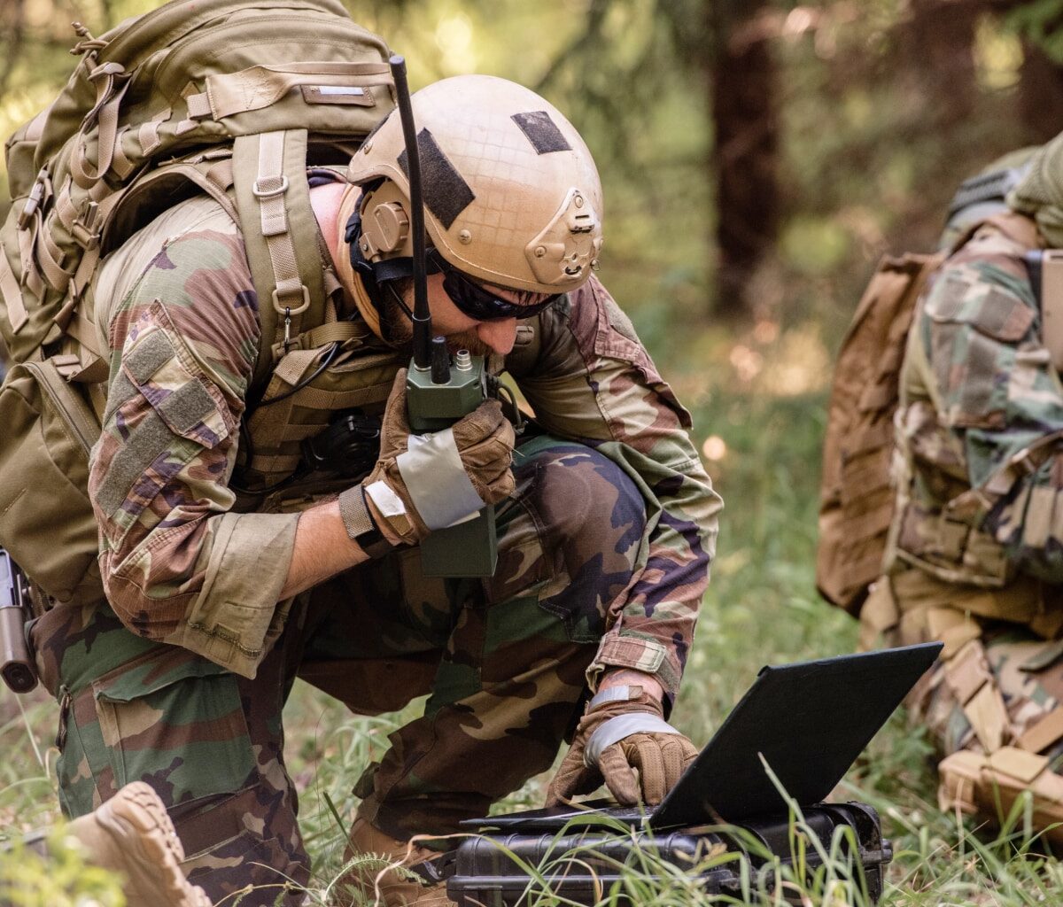 A soldier in camouflage gear and a helmet kneels in the grass, speaking into a radio and using a laptop, while another armed soldier sits nearby in a wooded area.