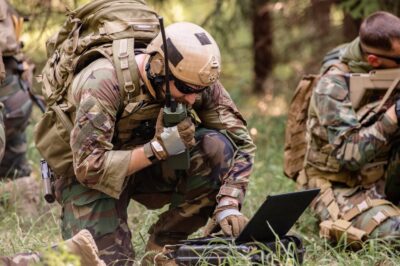 A soldier in camouflage gear and a helmet kneels in the grass, speaking into a radio and using a laptop, while another armed soldier sits nearby in a wooded area.