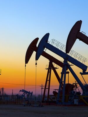 Oil pumpjacks and machinery silhouetted against a colorful sunset sky, with orange, yellow, and blue hues in the background.