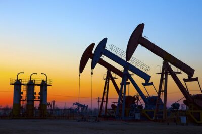 Oil pumpjacks and machinery silhouetted against a colorful sunset sky, with orange, yellow, and blue hues in the background.