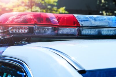 Close-up of red and blue emergency lights on top of a police car, with sunlight and trees blurred in the background.
