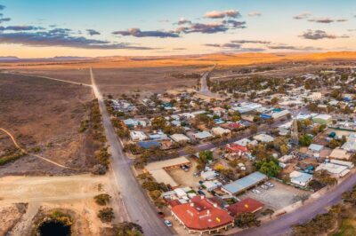 Aerial view of a small rural town surrounded by arid landscape, scattered houses, roads, and sparse vegetation, under a partly cloudy sky at sunset.