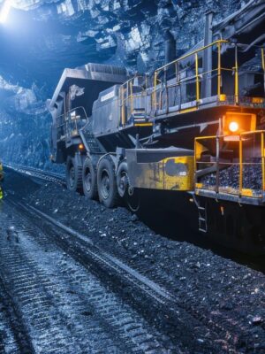 A worker in high-visibility clothing walks through a dimly lit underground mine tunnel beside large mining equipment, with rugged rock walls and tracks on the ground.