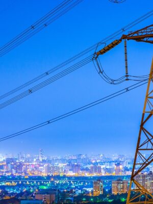 A large steel electricity pylon with power lines is seen in the foreground, overlooking a brightly lit cityscape at dusk with numerous buildings and a blue sky.