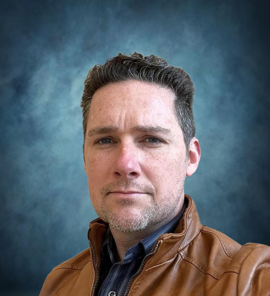 A man with short brown hair and light stubble wears a brown leather jacket over a collared shirt, looking directly at the camera. The background is a blue-gray gradient.