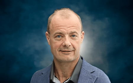 A middle-aged man with short, light hair wearing a blue blazer and dark shirt poses in front of a dark blue, cloudy background.