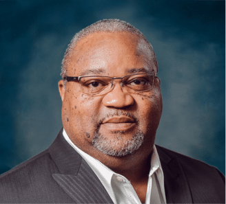 A middle-aged man with short gray hair, glasses, and a closely trimmed beard wears a dark suit jacket and white shirt, posing against a dark blue background.