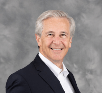 Smiling older man with gray hair wearing a dark suit jacket and white shirt, posing in front of a gray, softly blurred background.