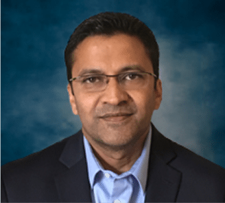 A man wearing glasses, a dark suit jacket, and a light blue shirt, poses in front of a blue and gray gradient background.