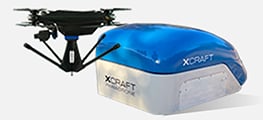 A black drone with four propellers sits next to a blue and white rectangular docking station labeled "XDRAFT".