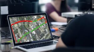 A person sits at a desk looking at a laptop displaying a map with a satellite view and a route marked in red, yellow, and green, possibly indicating traffic conditions or a planned path.
