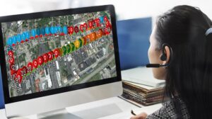 A woman wearing a headset looks at a computer screen displaying a street map with multicolored numbered markers overlaid, indicating various locations or tracking data points.