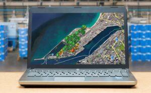 A laptop displays a satellite map with colored markers tracking locations at a port. Blurred storage racks and boxes are visible in the background, suggesting a warehouse setting.