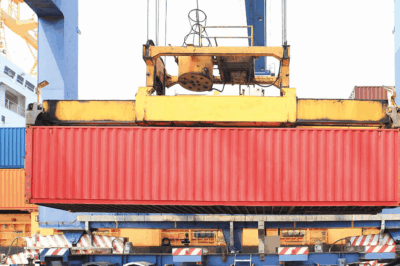 A large red shipping container is being lifted by a yellow crane at a shipping port, with stacked containers and part of a ship visible in the background.
