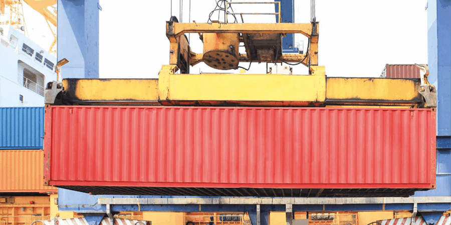 A large red shipping container is being lifted by a yellow crane at a shipping port, with stacked containers and part of a ship visible in the background.