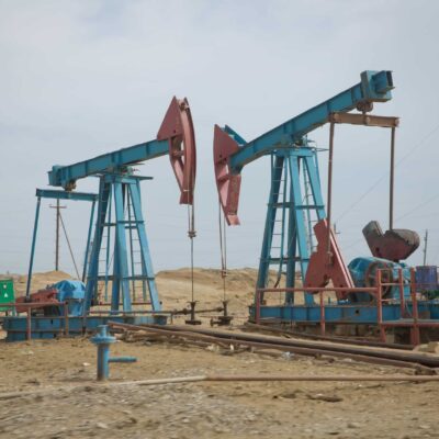 Two blue and red oil pumpjacks operate in a barren, sandy landscape with scattered industrial equipment and distant towers under a cloudy sky.