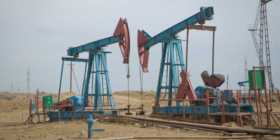 Two blue and red oil pumpjacks operate in a barren, sandy landscape with scattered industrial equipment and distant towers under a cloudy sky.
