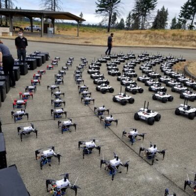 A large group of small robotic vehicles is arranged in organized rows outdoors on a paved area, with several people standing nearby. Trees and a shelter are visible in the background.