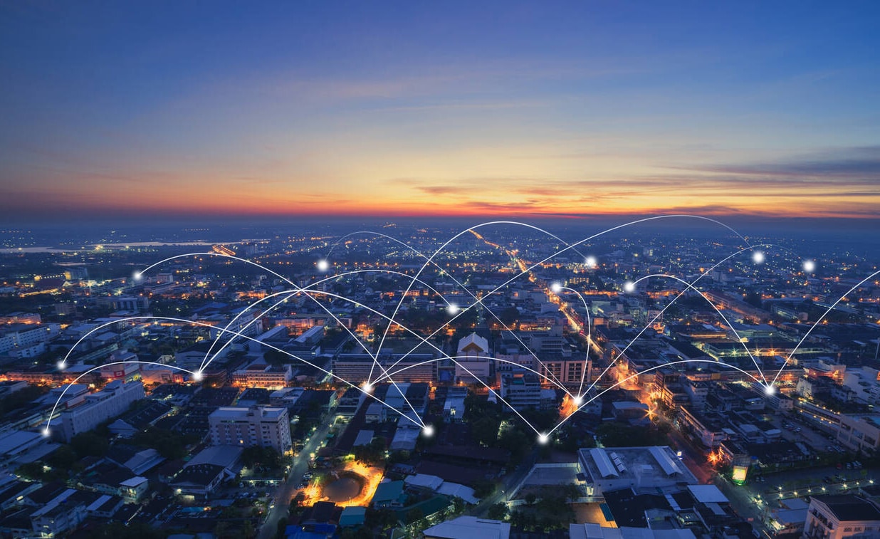 A cityscape at sunset with light trails and glowing points superimposed, symbolizing digital connectivity or a wireless network across the urban area.