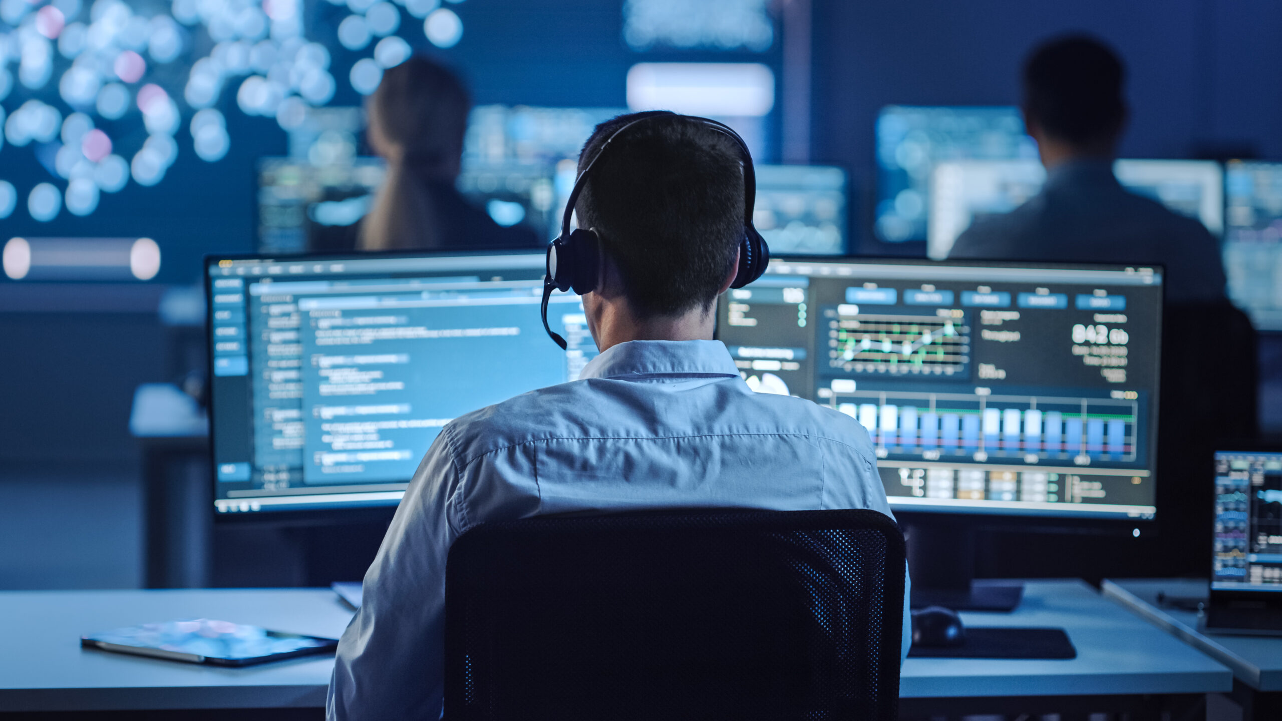 A person wearing a headset sits at a desk with multiple computer monitors displaying data, charts, and code in a dimly lit control room with other people working in the background.