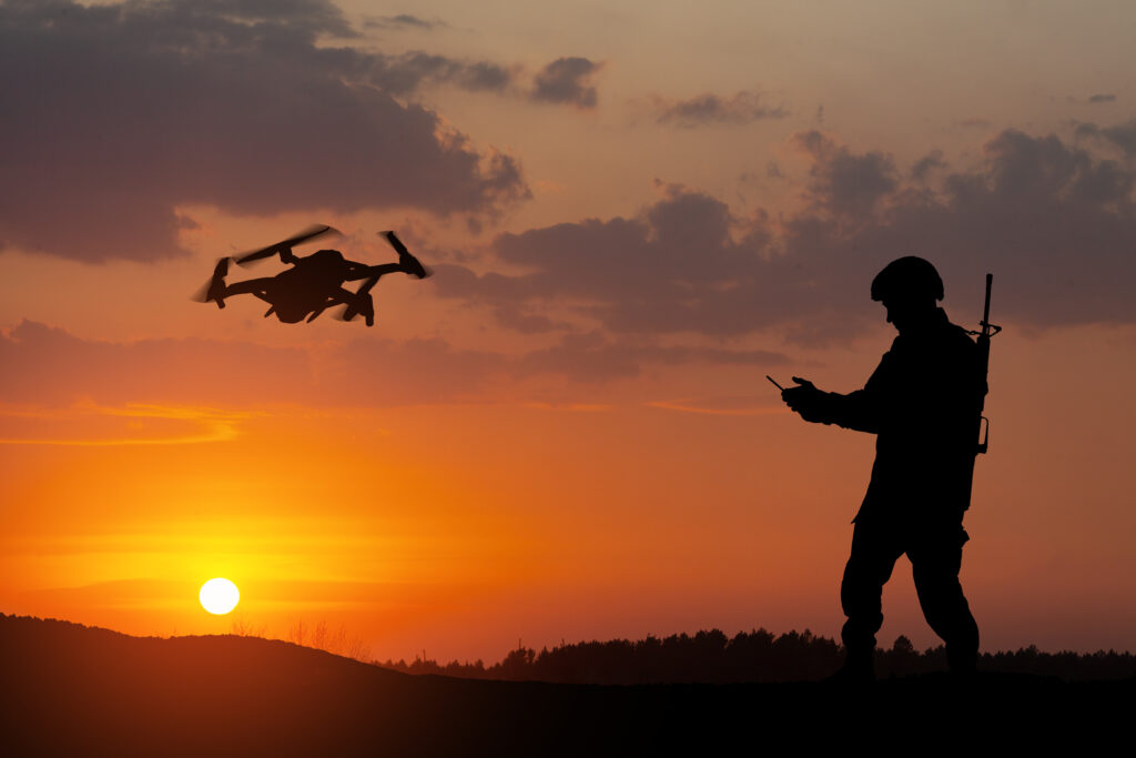 Silhouette of a soldier operating a drone at sunset, with the sun low on the horizon and clouds in the sky. The soldier holds a remote control, and a drone flies nearby.