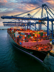 A large cargo ship loaded with multicolored shipping containers is docked at a port, with tall blue cranes ready for unloading at sunset. The sky is partly cloudy with warm tones reflecting on the water.