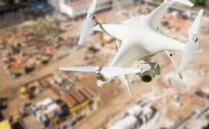 A white drone with a camera hovers above a construction site, capturing aerial footage of the area where equipment, materials, and workers are visible below.