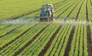 A tractor sprays pesticide or fertilizer over rows of green crops in a large, well-maintained agricultural field, demonstrating modern farming practices.