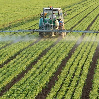 A tractor sprays pesticide or fertilizer over rows of green crops in a large, well-maintained agricultural field, demonstrating modern farming practices.