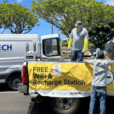 Two men stand by trucks in a parking lot, attaching a yellow banner that reads “FREE WiFi & Recharge Station.” The scene is sunny, with trees and a SITECH van visible in the background.