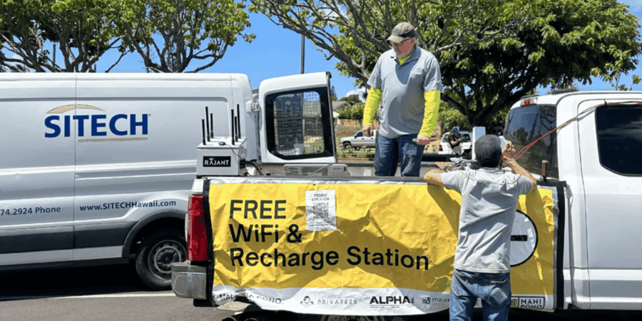 Two men stand by trucks in a parking lot, attaching a yellow banner that reads “FREE WiFi & Recharge Station.” The scene is sunny, with trees and a SITECH van visible in the background.