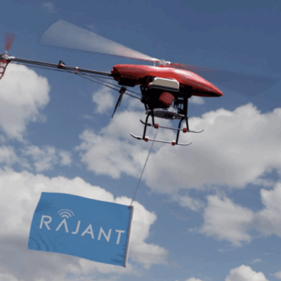 A red drone flies against a partly cloudy sky, carrying a sign with the blue Rajant logo and wireless signal icon hanging from its underside.
