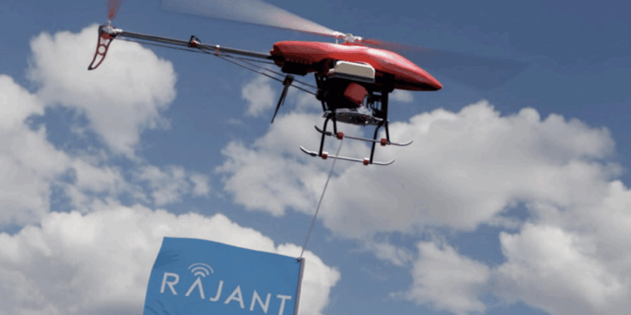 A red drone flies against a partly cloudy sky, carrying a sign with the blue Rajant logo and wireless signal icon hanging from its underside.