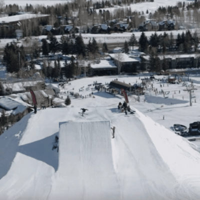Aerial view of a snowy ski resort with snowboarders performing tricks off a large jump, surrounded by trees, buildings, and people watching from below.