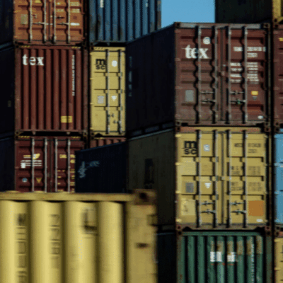 Stacks of colorful shipping containers are arranged in rows under a partly cloudy sky, indicating a busy shipping yard or port area.