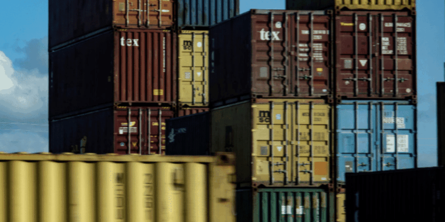 Stacks of colorful shipping containers are arranged in rows under a partly cloudy sky, indicating a busy shipping yard or port area.